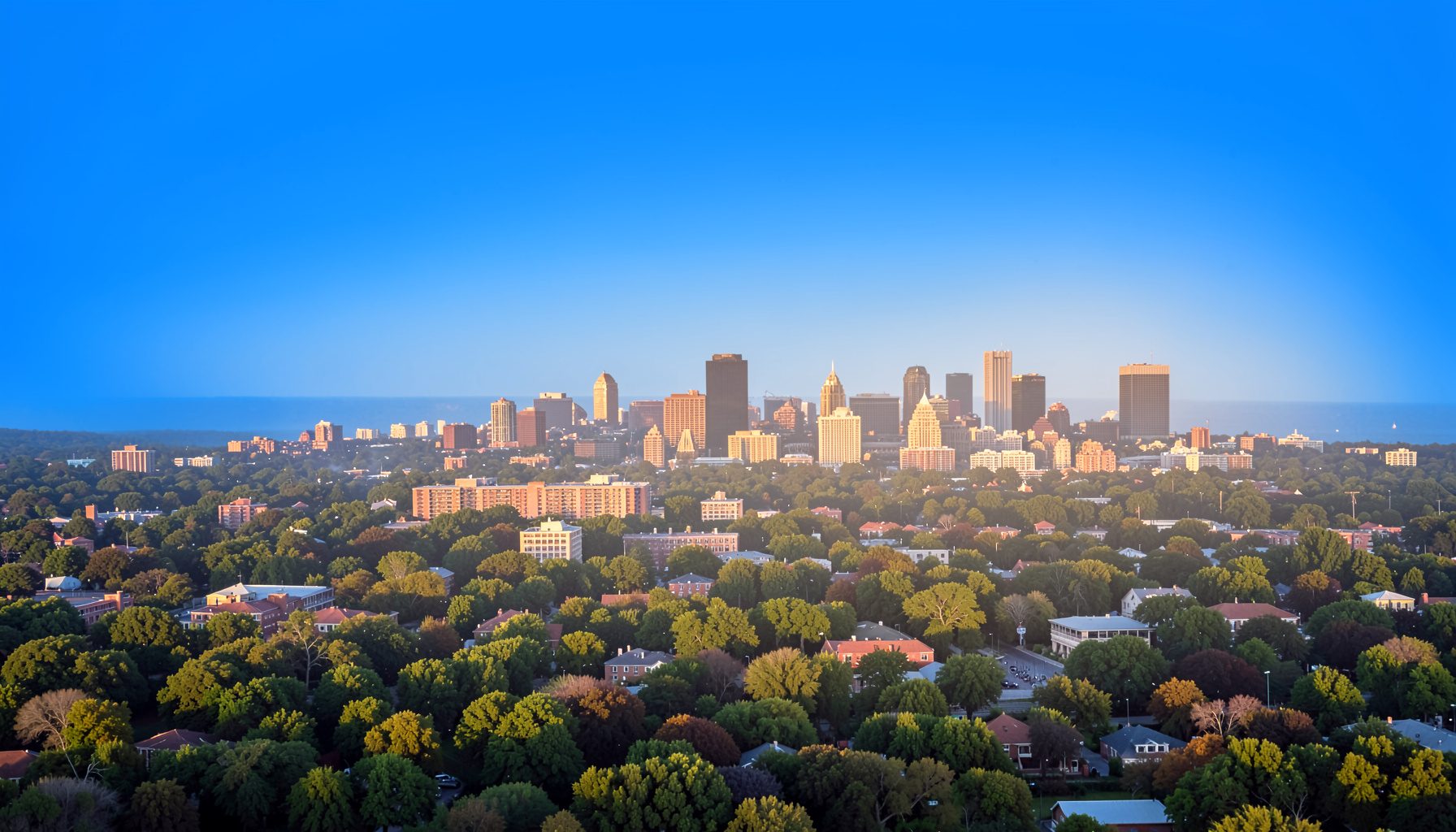 A wide aerial view of the Research Triangle skyline at golden hour, with soft haze over the tree canopy and downtown towers in the distance, warm amber and slate tones.