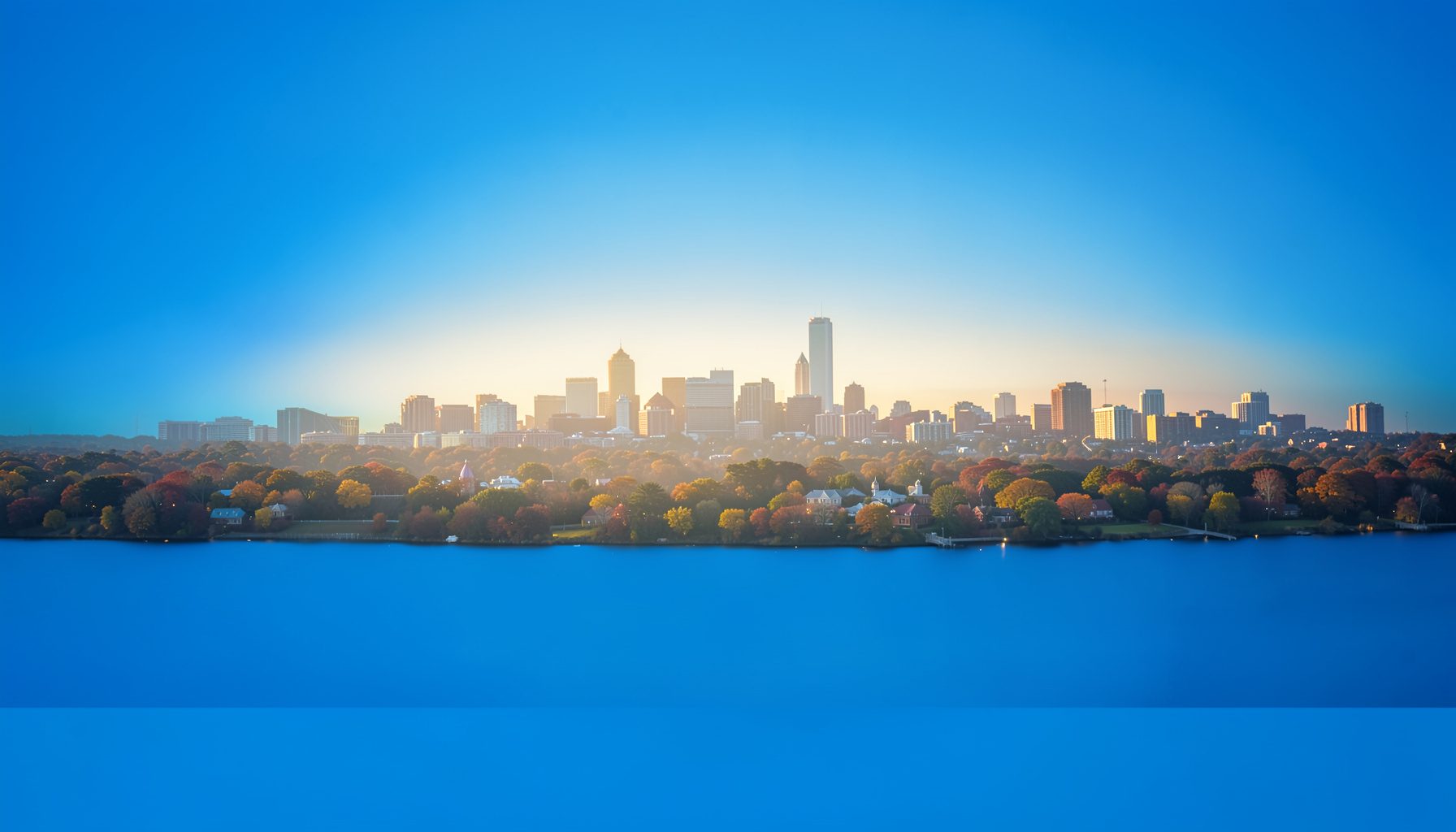 A wide landscape photograph of the Research Triangle skyline at golden hour, with soft warm light filtering through a light haze over the city, conveying a calm and community-focused atmosphere.